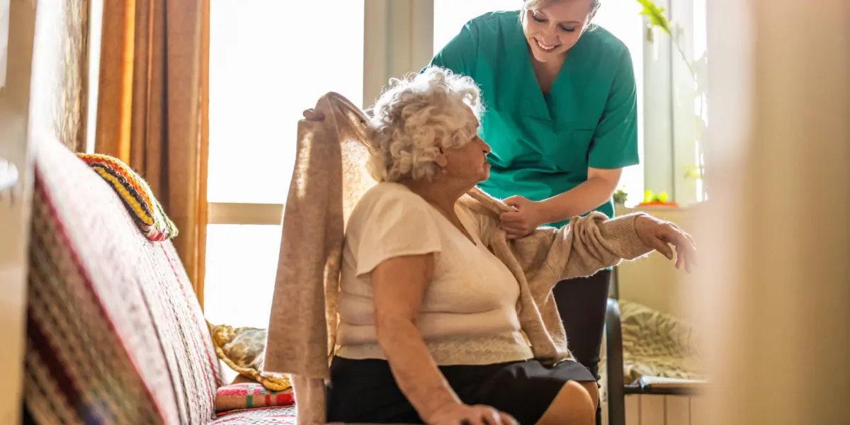 A caregiver offers support to an older adult seated near a window.