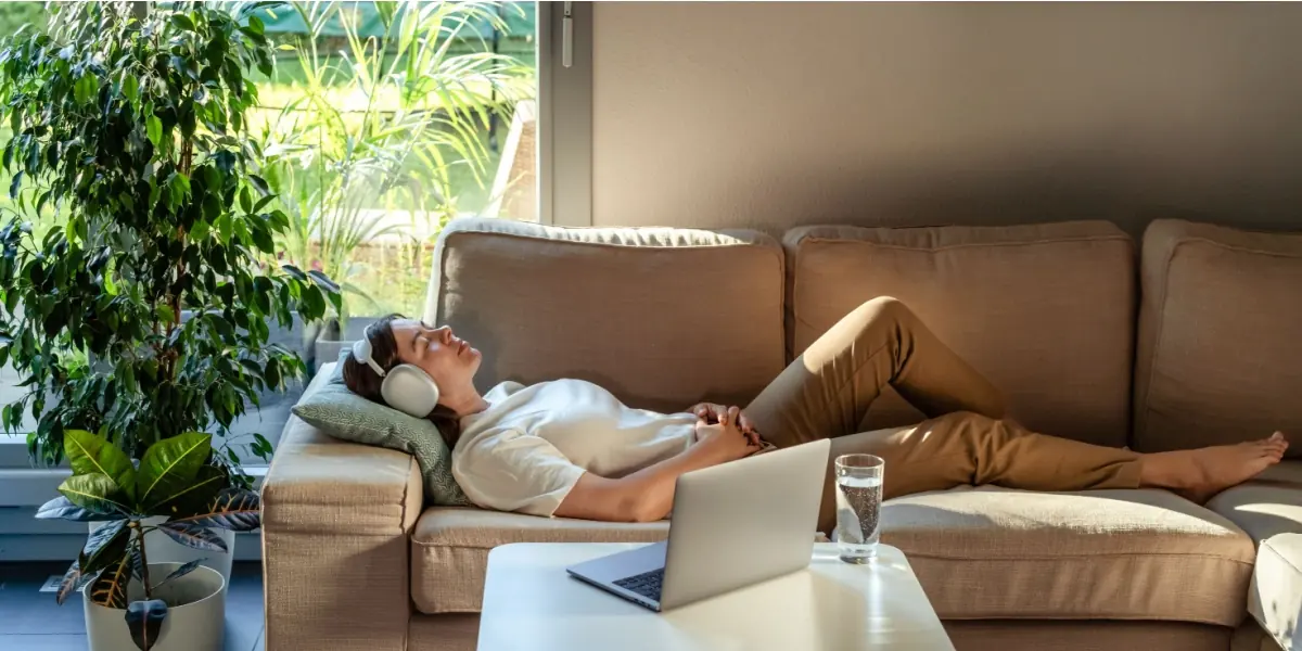 A person wearing headphones rests on a sofa beside plants.
