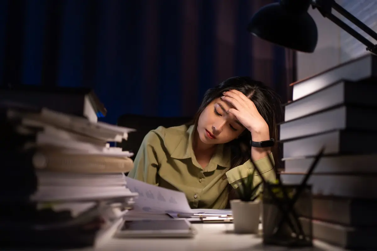 A person pauses at a desk surrounded by books and work.