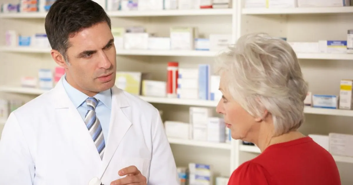 A pharmacist talks with an older adult at a pharmacy counter.