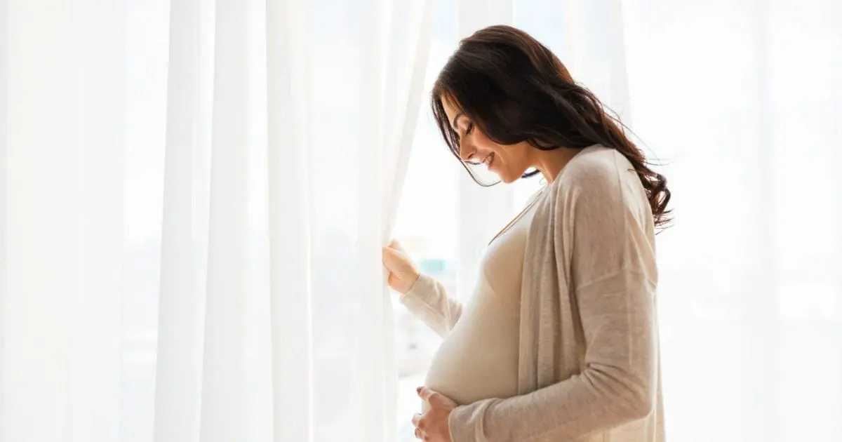 A pregnant woman stands beside a bright window.