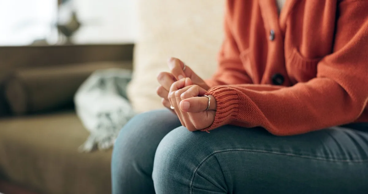 A person in a rust-colored sweater sits with hands clasped in their lap.