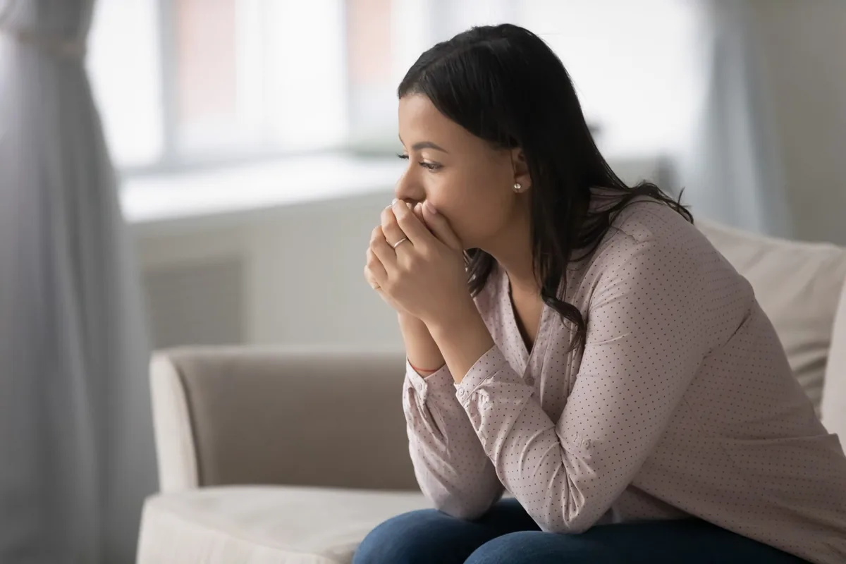 A woman sits thoughtfully on a sofa with her hands near her face.