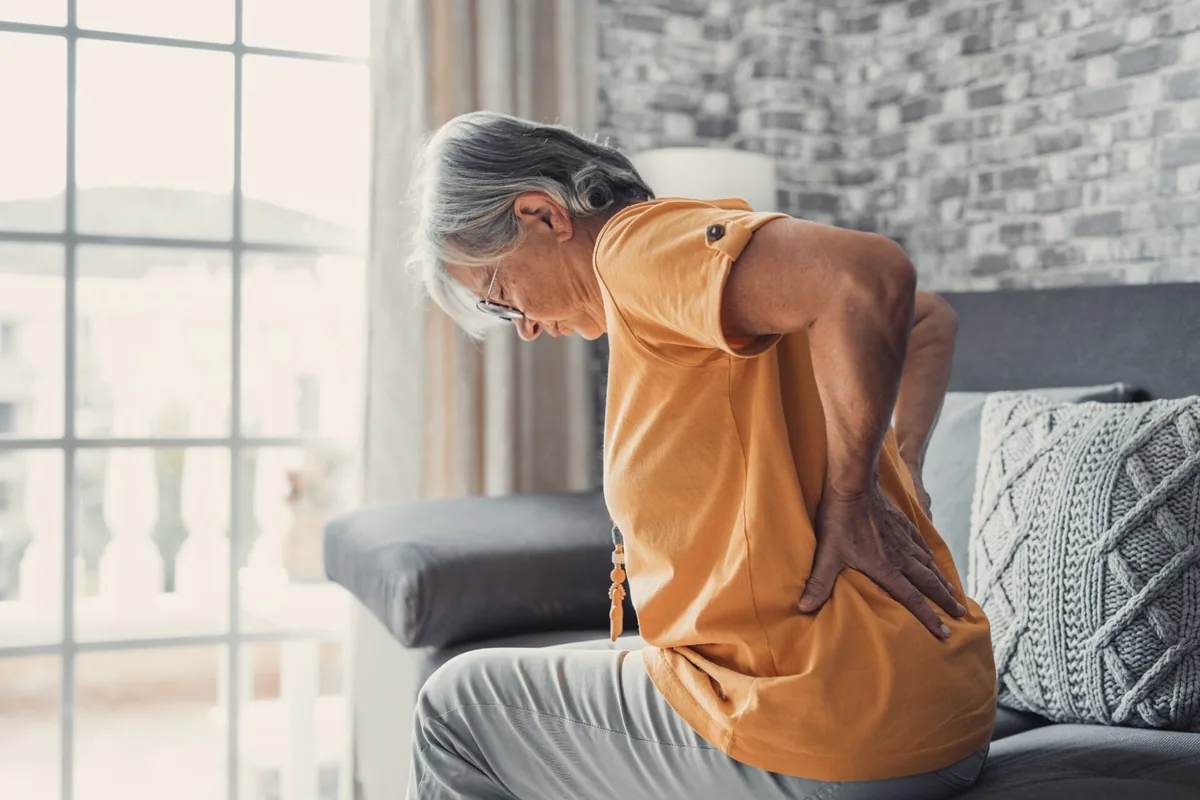 An older adult stretches while seated beside a bright window.