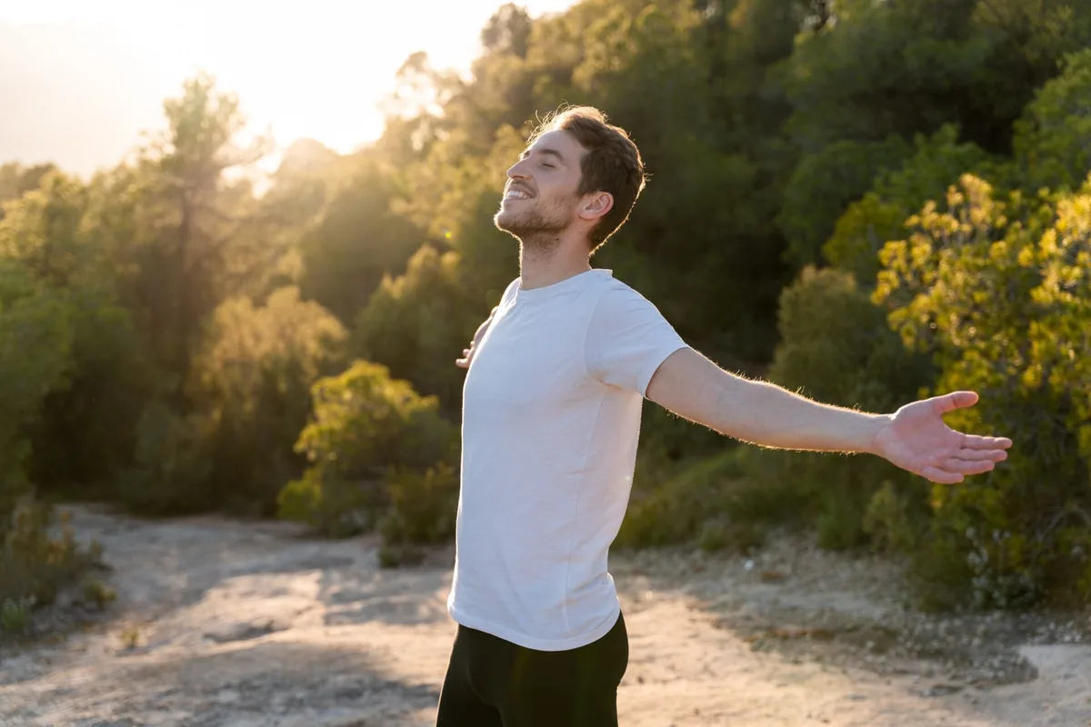 A man opens his arms while standing in a sunlit park.