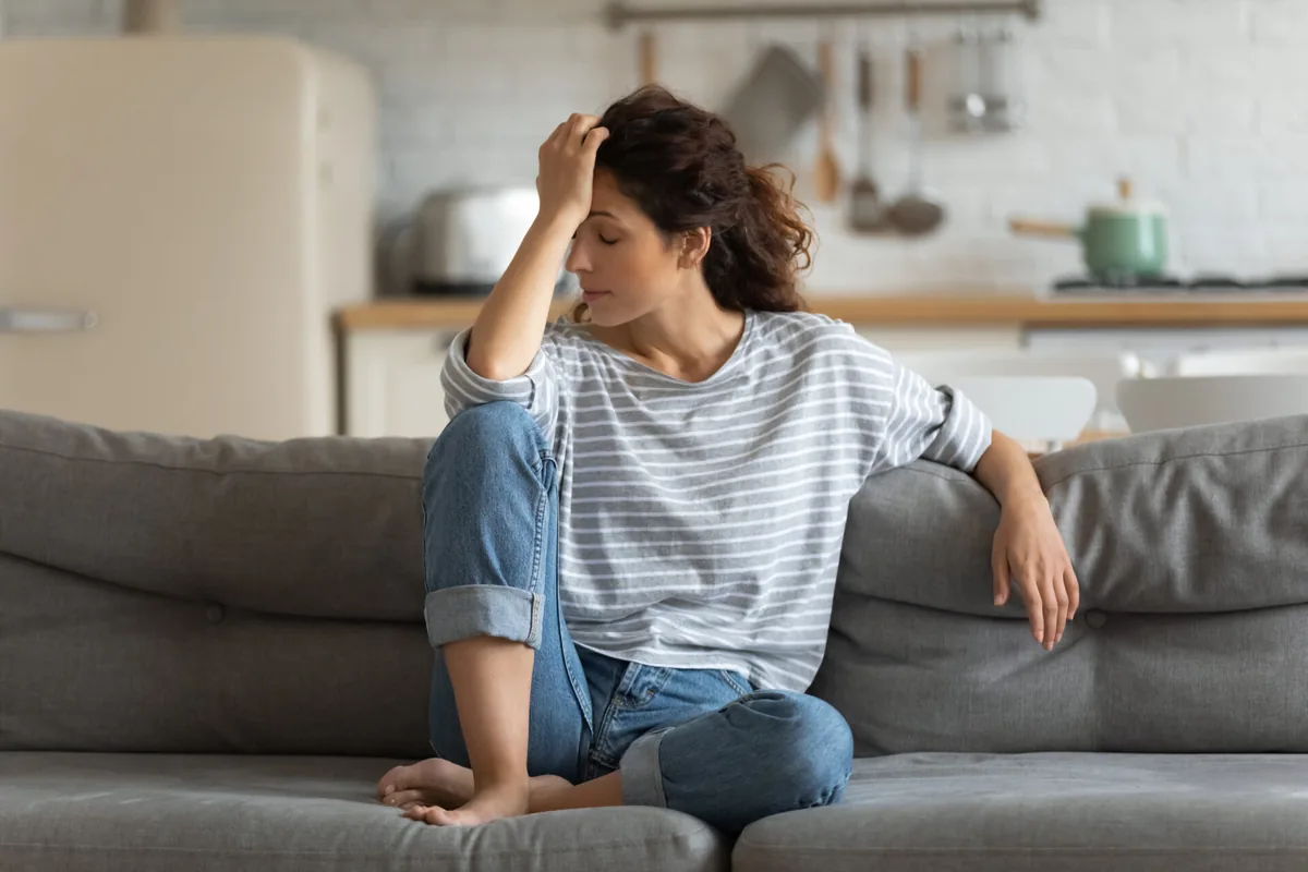A woman sits on a sofa and pauses with a hand resting against her forehead.