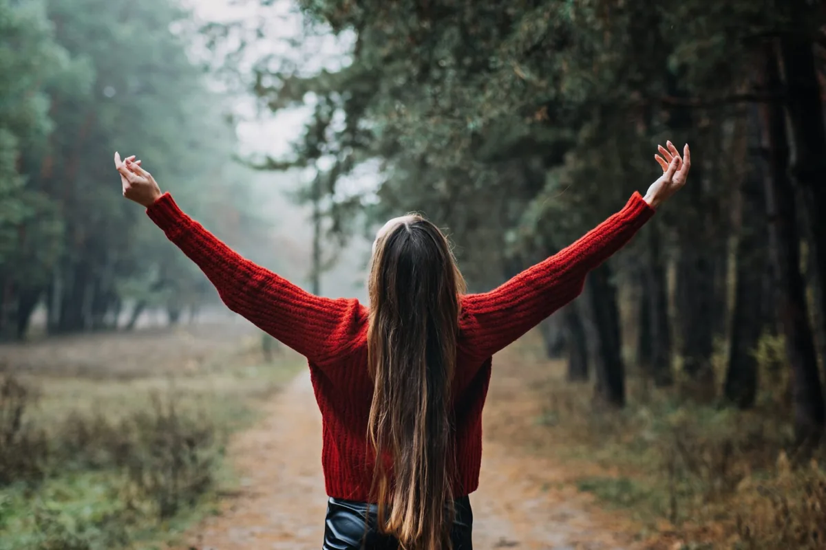 A person lifts their arms beneath tall trees in a green forest.