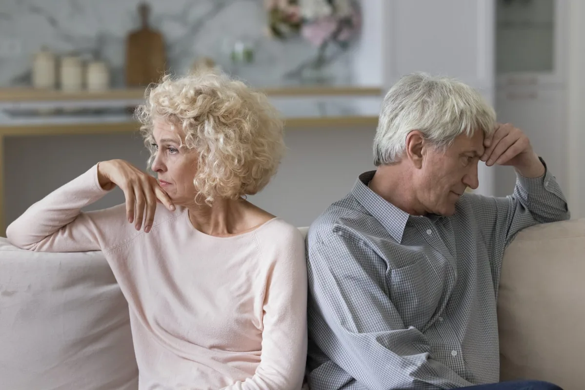 An older couple sits apart on a sofa, each looking thoughtful.