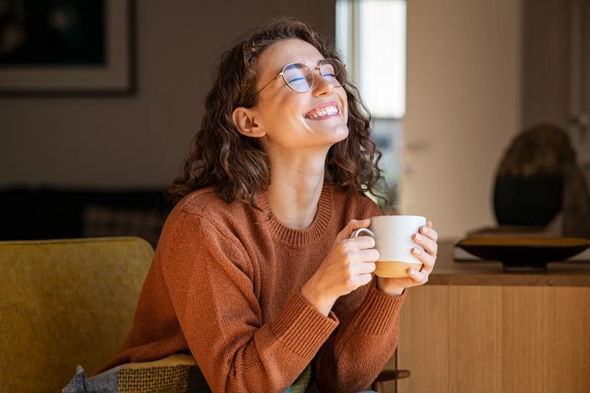 A woman smiles while holding a warm mug in a cozy room.
