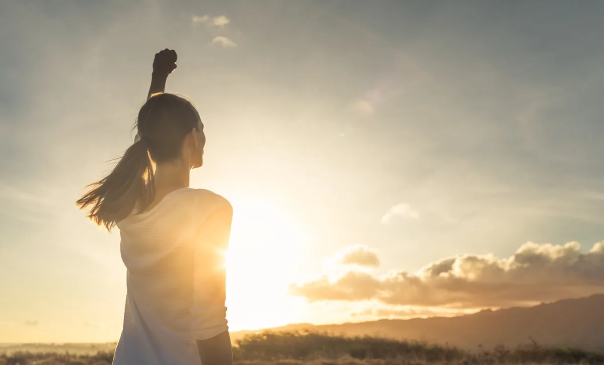 A person stands on a hill with one arm raised toward the sunrise.