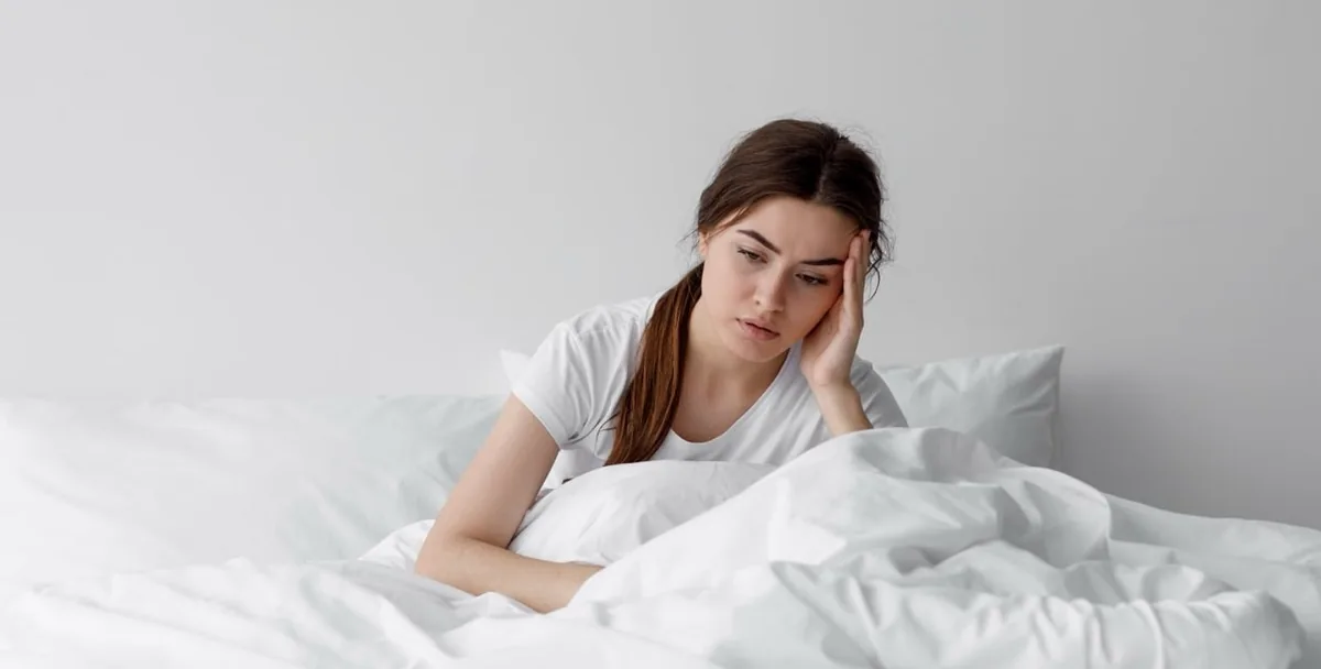 A woman sits quietly in bed with a duvet across her lap.