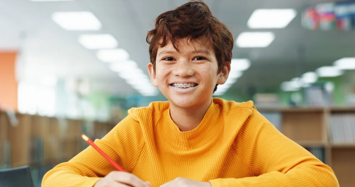 A young person smiles while writing at a classroom desk.