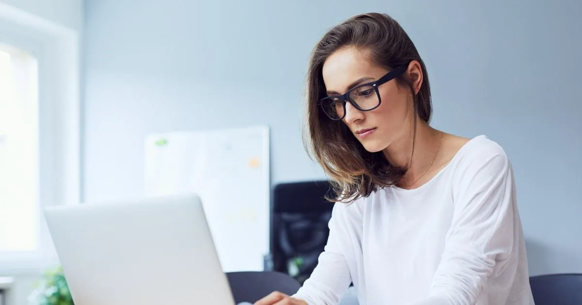 A woman wearing glasses works on a laptop in a bright room.