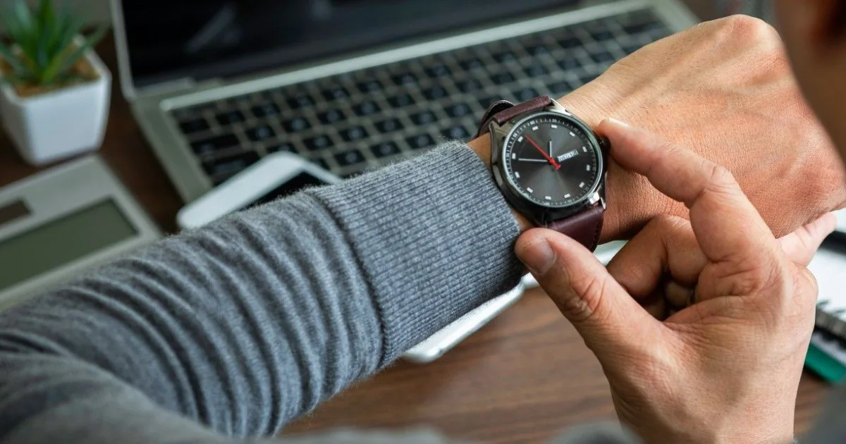 A person checks a wristwatch beside an open laptop.