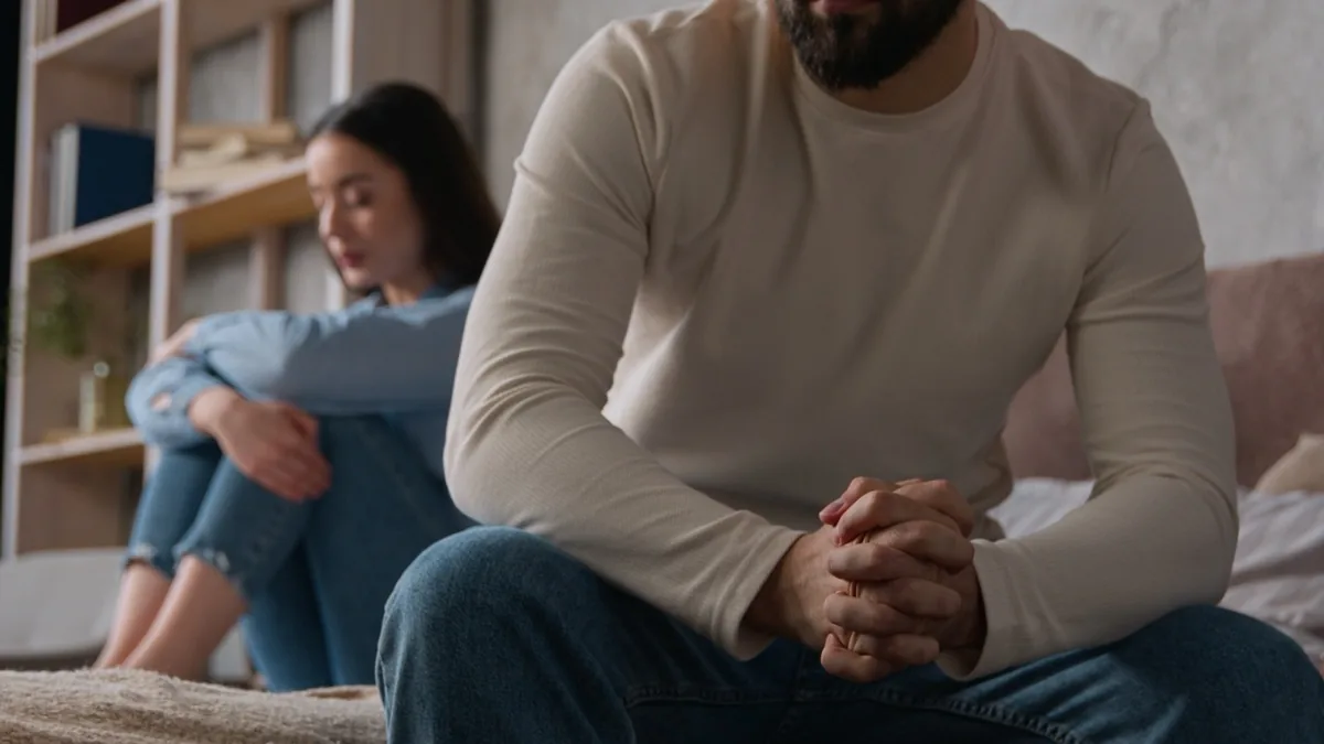 A couple sits apart on a sofa, each looking thoughtful.