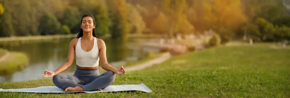 A woman sits cross-legged beside a lake in a green park.