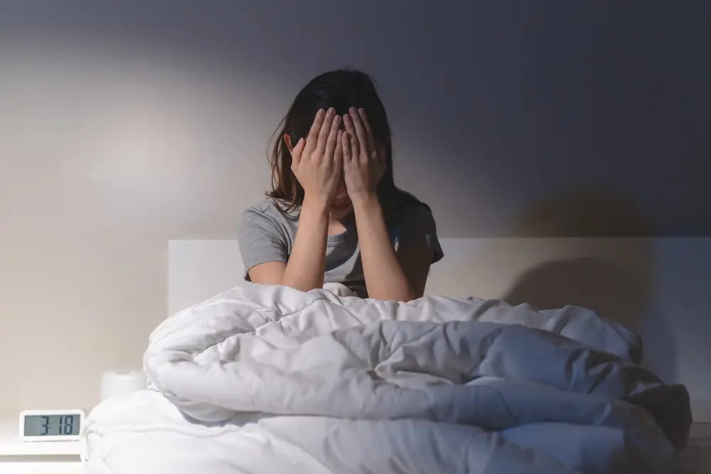 A woman sits on a bed with her hands near her face.