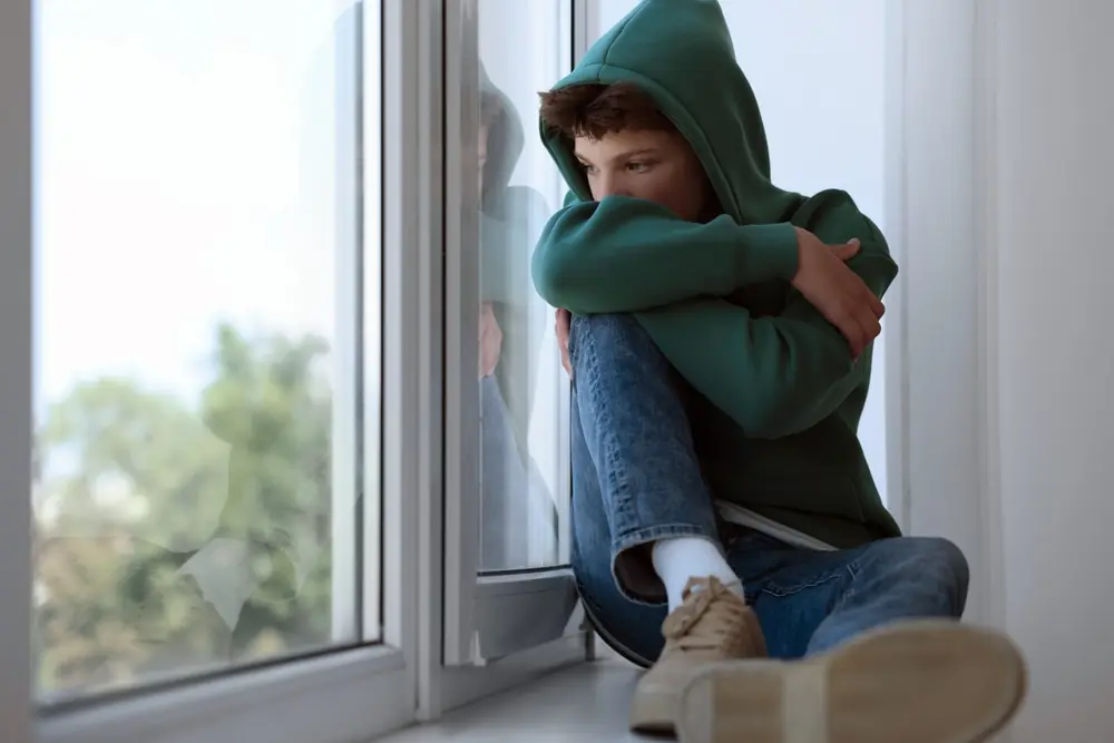 A young person sits beside a window with their knees drawn up.