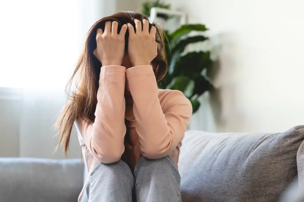 A woman rests her face in her hands while seated on a sofa.