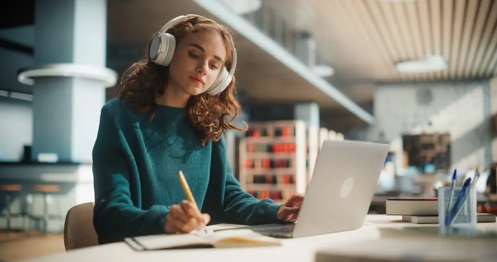 A woman writes in a notebook beside a laptop.